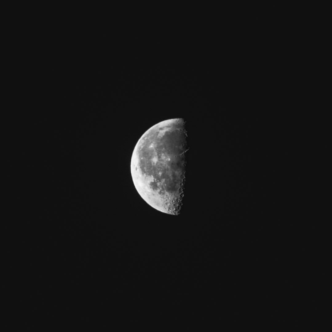 Detailed monochrome shot of the moon in partial phase against a stark black sky. The moon's surface is textured and cratered.