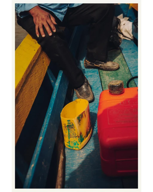 Man sits on a colorful boat with blue, yellow, and red elements, wearing black pants, a blue shirt, and dark shoes next to plastic containers.