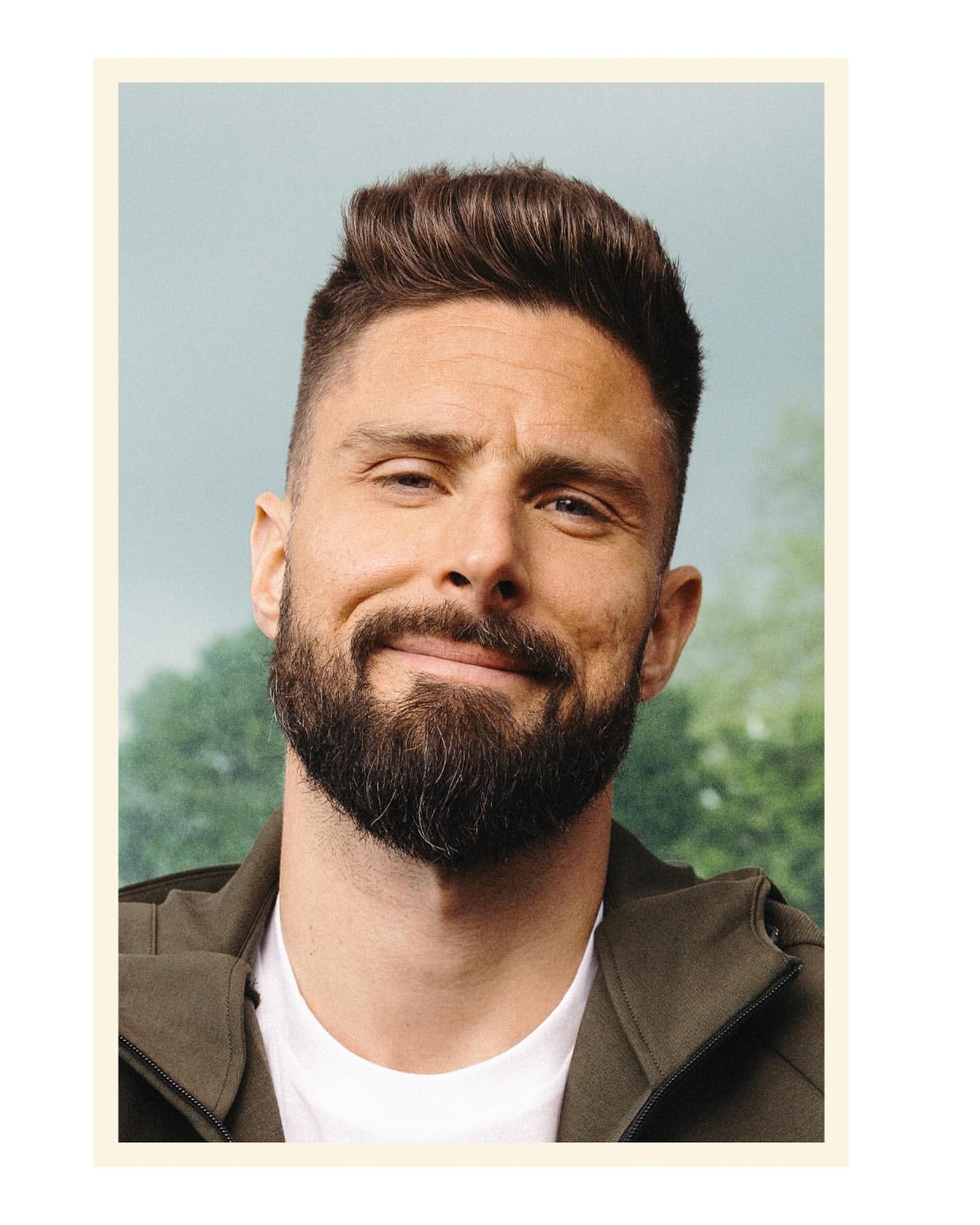 Headshot of male athlete with dark hair and a beard, wearing a dark green jacket, set against a soft, blurred background.