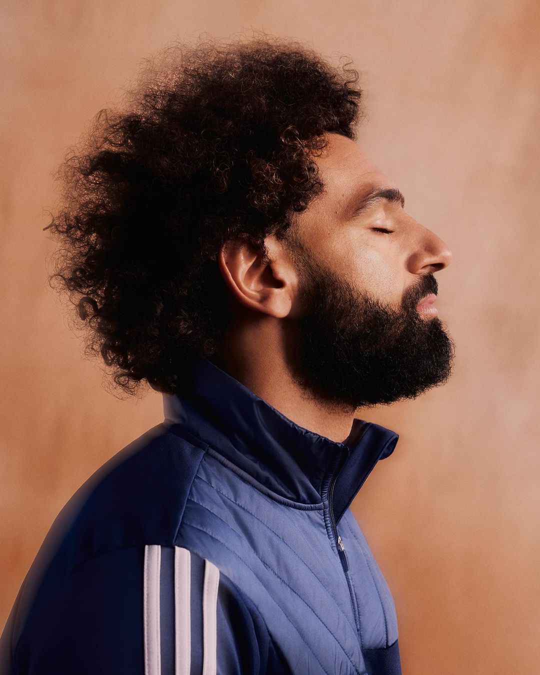 Portrait of a bearded man with curly hair wearing a blue Adidas track jacket against an orange background with closed eyes.