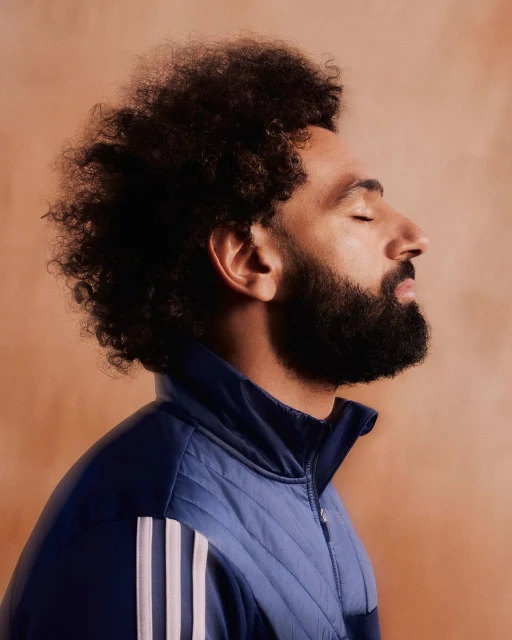 Portrait of a bearded man with curly hair wearing a blue Adidas track jacket against an orange background with closed eyes.