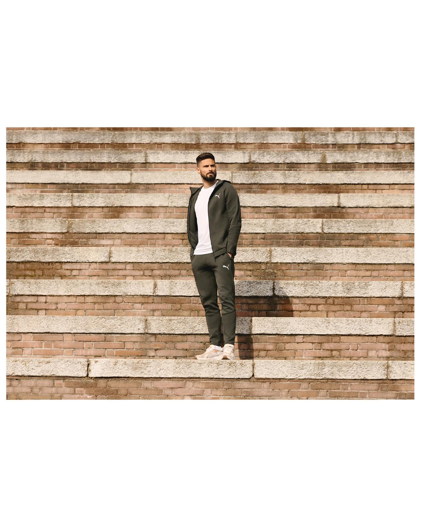 Adult male wearing a dark green tracksuit standing on a stone staircase in an outdoor setting on a sunny day.