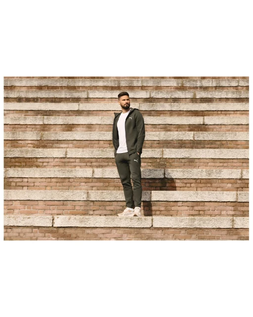 Adult male wearing a dark green tracksuit standing on a stone staircase in an outdoor setting on a sunny day.