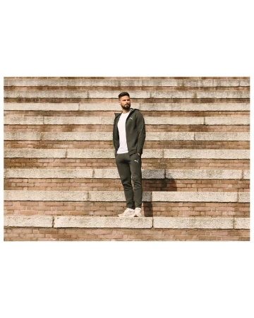Adult male wearing a dark green tracksuit standing on a stone staircase in an outdoor setting on a sunny day.