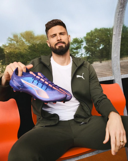 An adult male poses outside, displaying a purple and pink Puma soccer shoe while sitting on a bench, wearing an athletic jacket and pants.