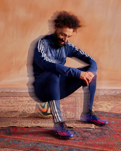 An athlete in sports clothing sits on a soccer ball on a patterned rug in a studio setting with an orange-toned background.