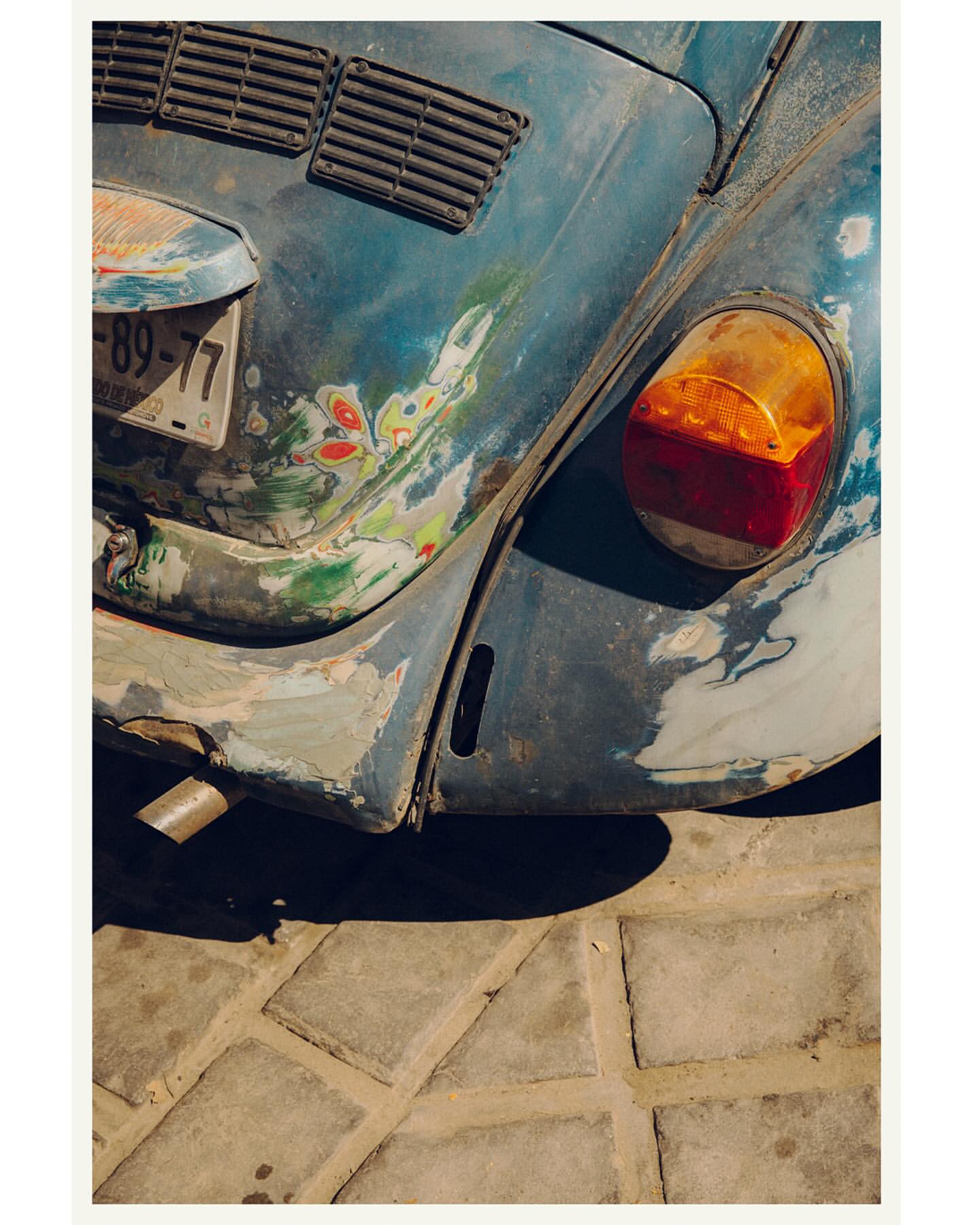 Close-up of a distressed blue Volkswagen Beetle with a visible taillight, license plate, and paint damage, parked on a cobblestone surface.