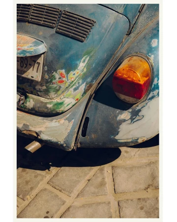 Close-up of a distressed blue Volkswagen Beetle with a visible taillight, license plate, and paint damage, parked on a cobblestone surface.