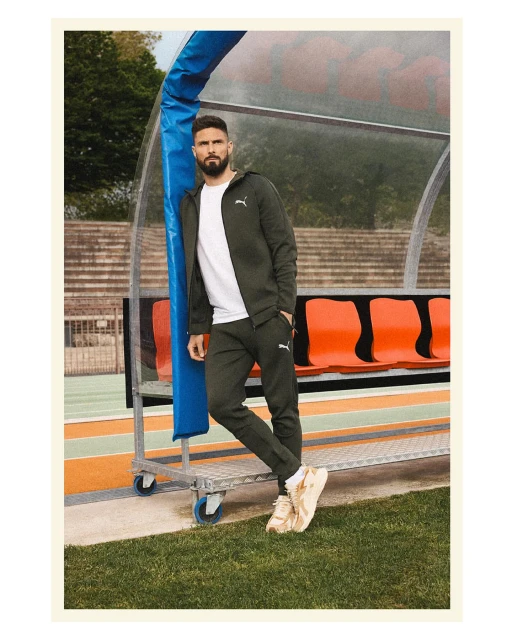 Man in athletic jacket and pants posing next to stadium seating on a sunny day with green grass in foreground, wearing sneakers.