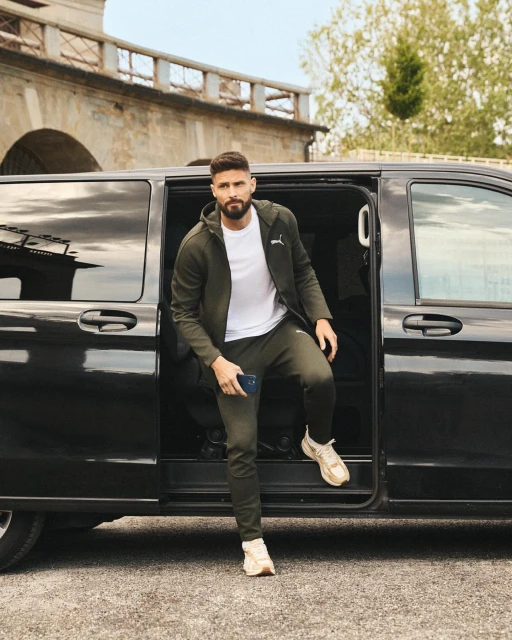 A man in a green tracksuit exits a black van, wearing a white t-shirt and sneakers, with an arched stone bridge in the background.