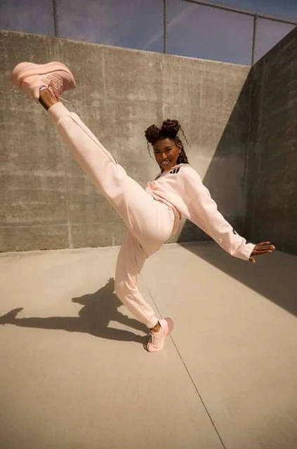 A woman in pink athleisure wear strikes a dynamic pose with one leg raised high, set against a gray concrete wall under natural light.