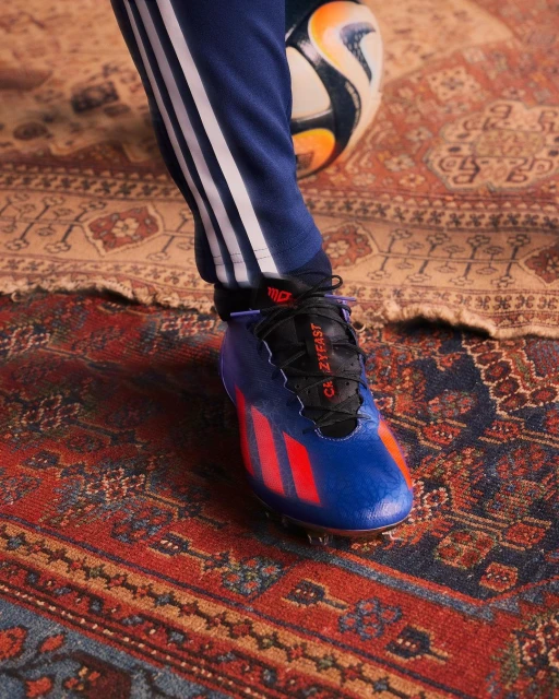 Close-up of a blue and orange Adidas soccer cleat on a red patterned rug, with a soccer ball and striped pants visible.