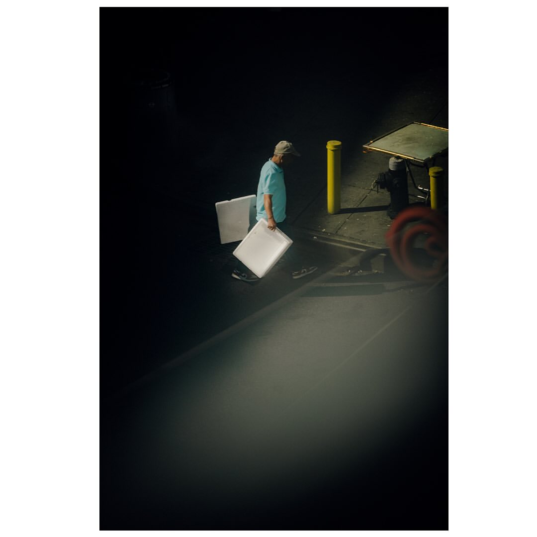 Man in a blue shirt carries two white boards in a dark outdoor setting near a cart with blurred wheels and a yellow bollard.