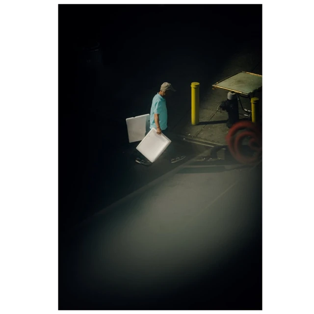 Man in a blue shirt carries two white boards in a dark outdoor setting near a cart with blurred wheels and a yellow bollard.