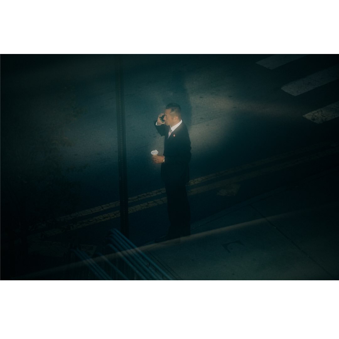 Man in a suit on a city street, drinking from a white paper cup while talking on his cell phone under street lights at night.