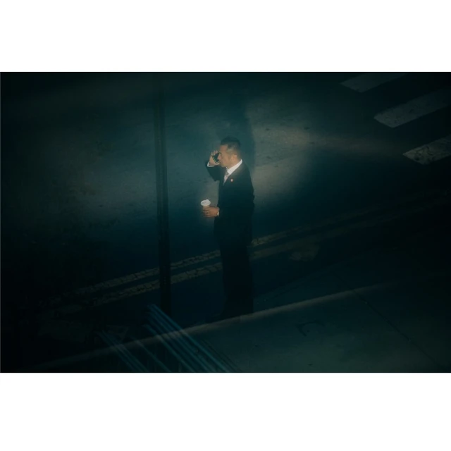Man in a suit on a city street, drinking from a white paper cup while talking on his cell phone under street lights at night.