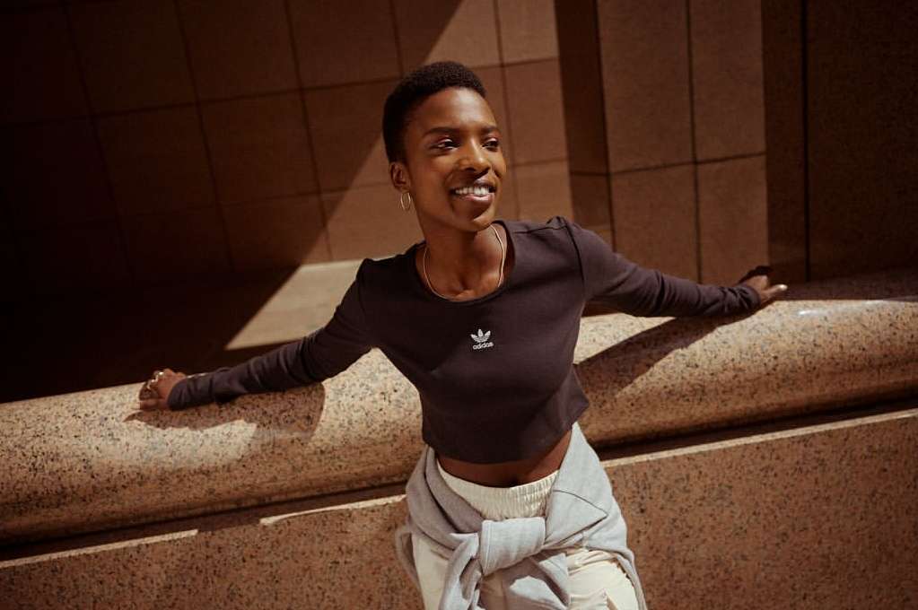 A smiling Black woman poses outdoors against a textured wall in a cropped black long-sleeved shirt, with a sweatshirt tied around her waist.