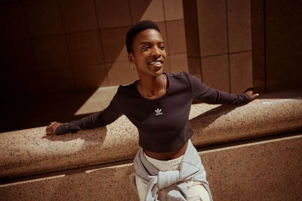 A smiling Black woman poses outdoors against a textured wall in a cropped black long-sleeved shirt, with a sweatshirt tied around her waist.