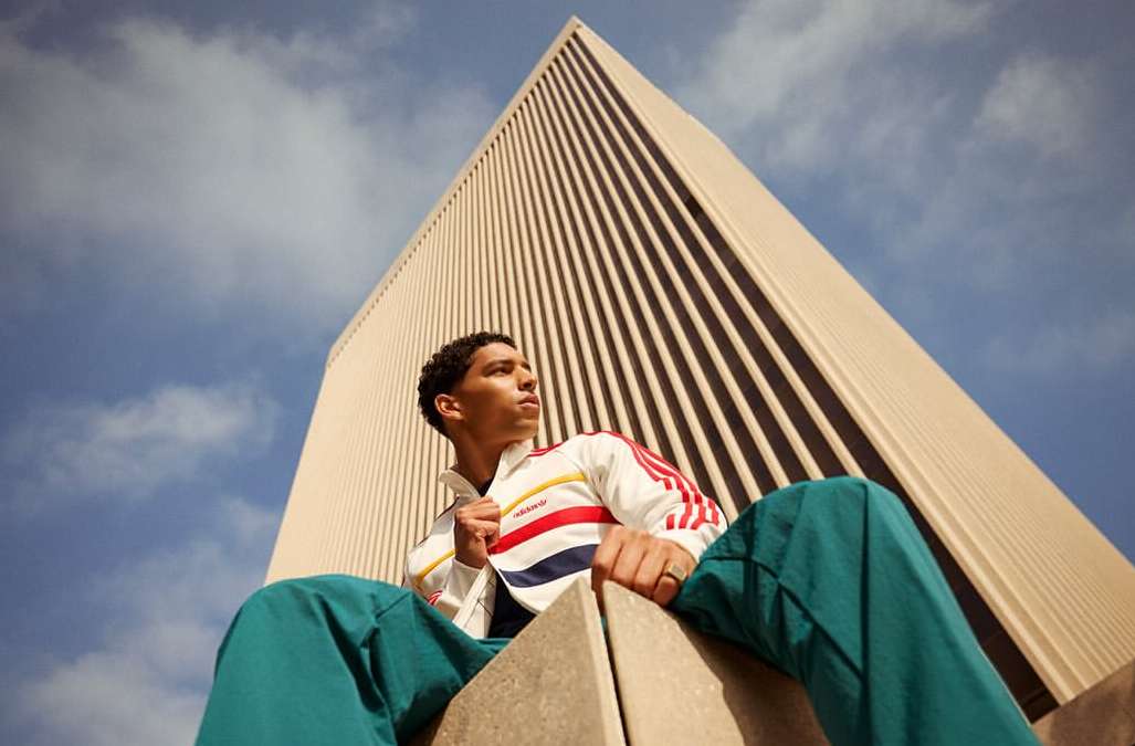 Adult male in a colorful jacket poses in front of an urban building and a blue sky with clouds in an eye-level outdoor shot.