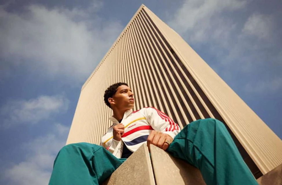 Adult male in a colorful jacket poses in front of an urban building and a blue sky with clouds in an eye-level outdoor shot.