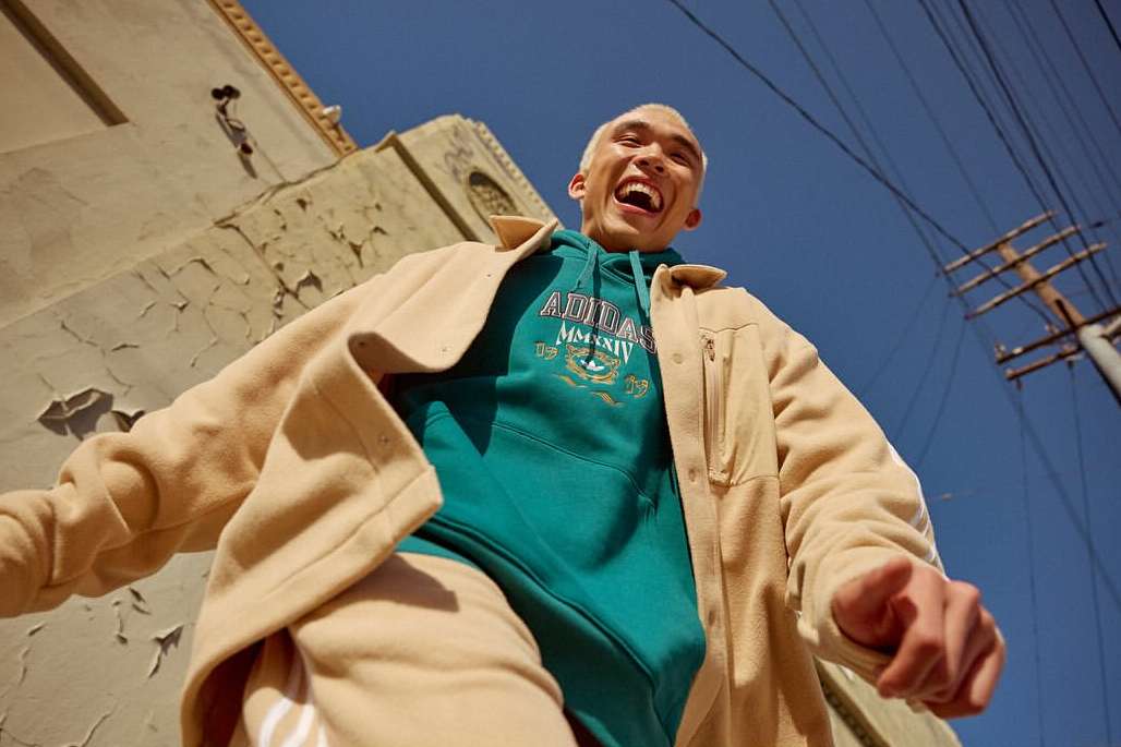 A young adult smiles widely while wearing a teal Adidas hoodie and tan overshirt in an urban setting under a clear blue sky.