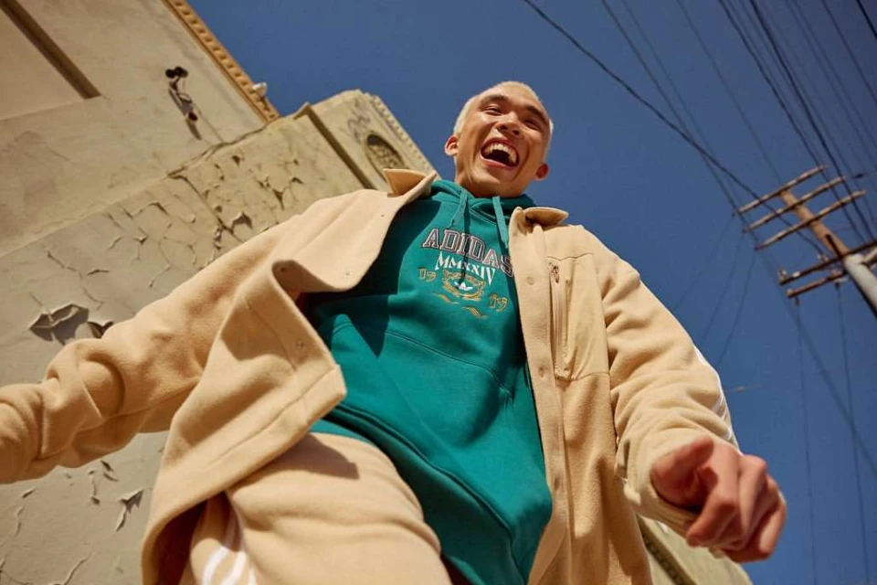A young adult smiles widely while wearing a teal Adidas hoodie and tan overshirt in an urban setting under a clear blue sky.