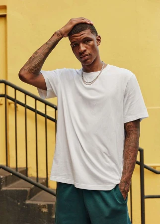 Black man with tattoos wearing a white t-shirt and green trousers standing outdoors near stairs against a yellow wall