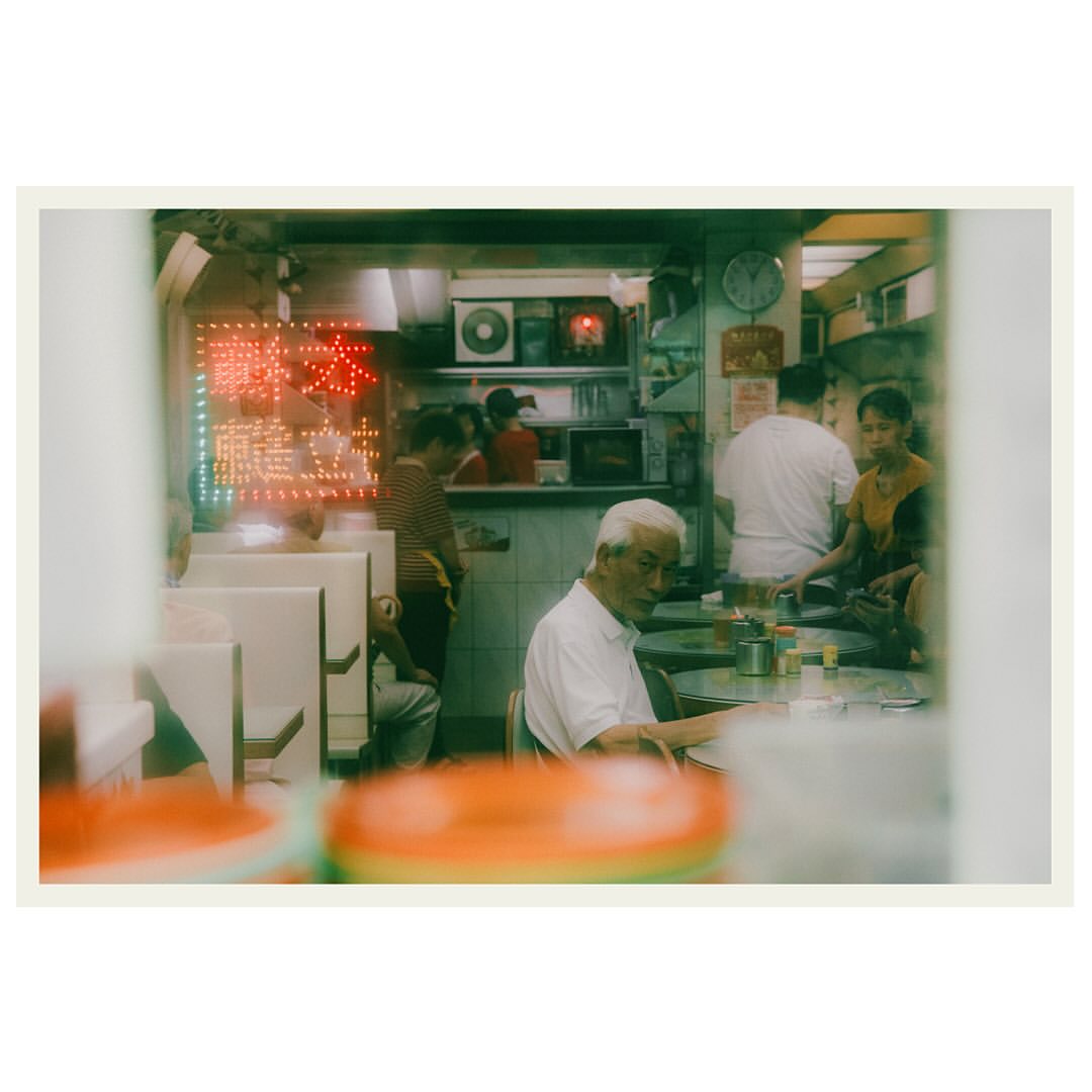 Elderly Asian man sits at a table in a restaurant, other people are visible in background, neon sign hangs in the background.