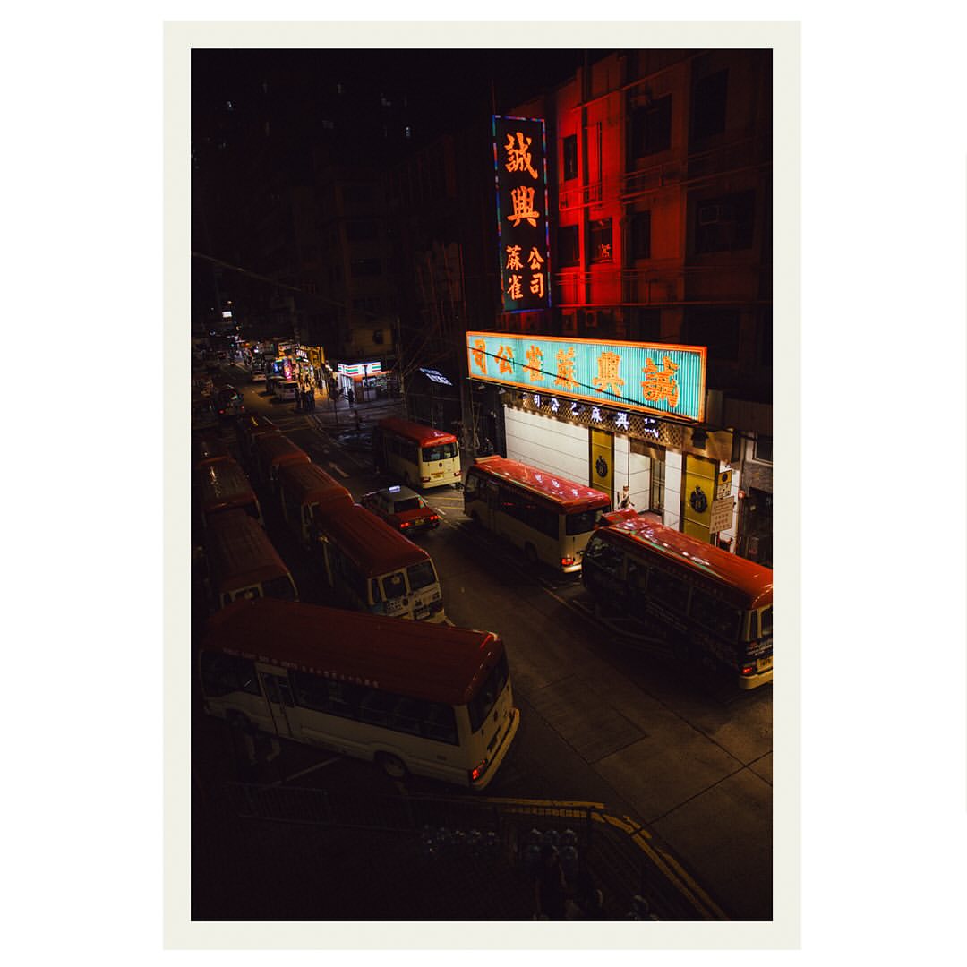 Night scene in an urban environment featuring several red and white mini-buses parked on a street, lit by bright commercial signage.