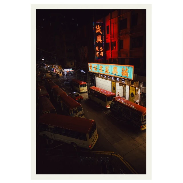 Night scene in an urban environment featuring several red and white mini-buses parked on a street, lit by bright commercial signage.