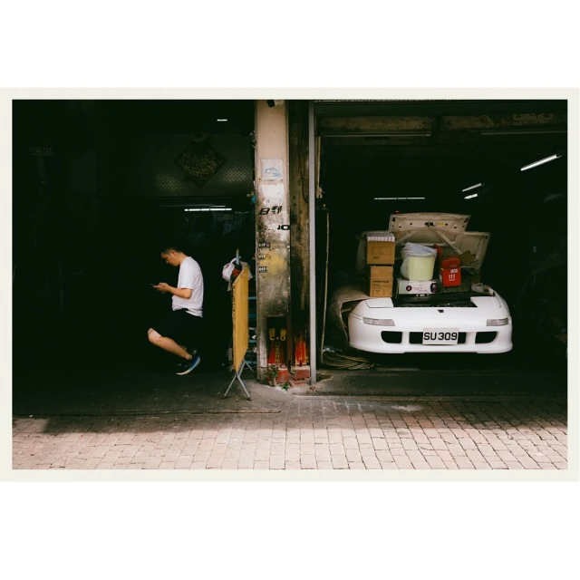 Man crouching and using a phone near a white car parked inside a garage with boxes and items inside of the vehicle.