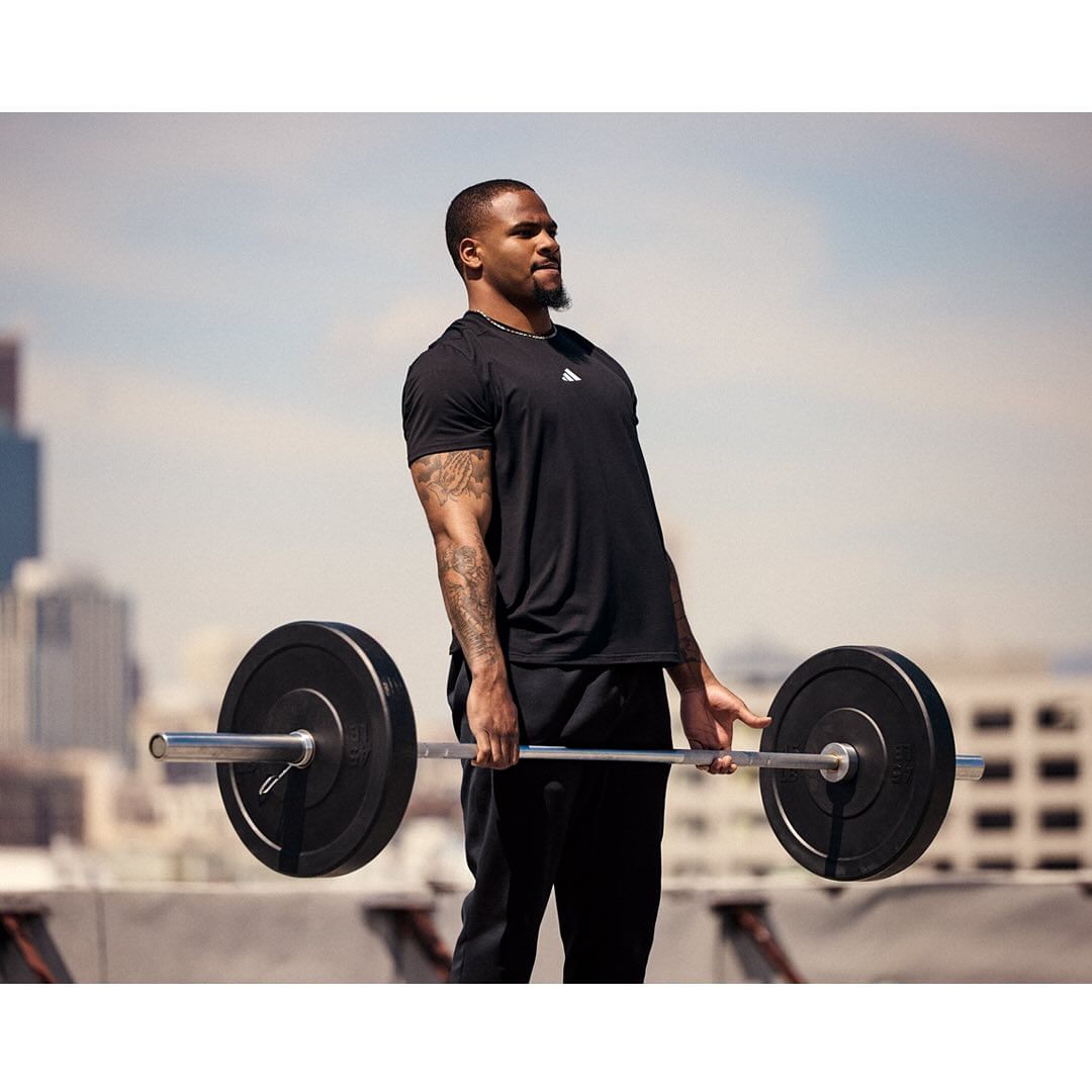 Athletic man in black t-shirt exercising with a barbell on a rooftop with city buildings in the background on a sunny day.