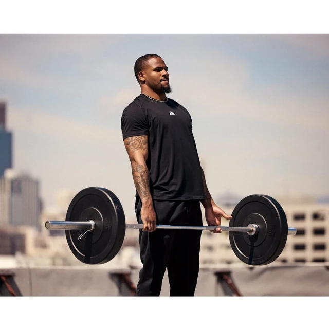 Athletic man in black t-shirt exercising with a barbell on a rooftop with city buildings in the background on a sunny day.
