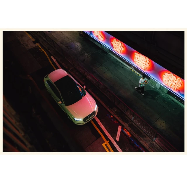 Overhead view of white Audi car on dark urban street at night with pedestrian walking along bright neon sign.