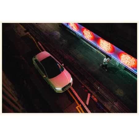 Overhead view of white Audi car on dark urban street at night with pedestrian walking along bright neon sign.