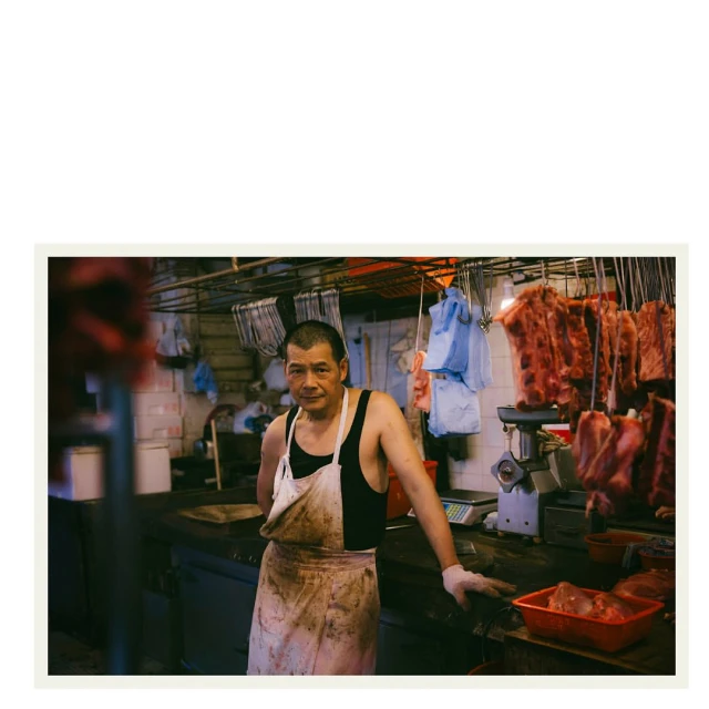 Asian man wearing a dirty apron in an indoor food market with hanging meat and kitchen equipment in the background, medium shot.