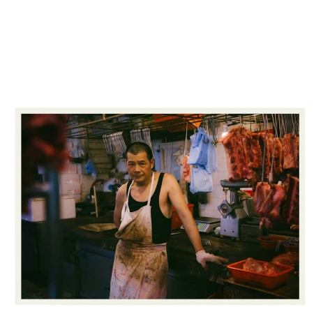 Asian man wearing a dirty apron in an indoor food market with hanging meat and kitchen equipment in the background, medium shot.