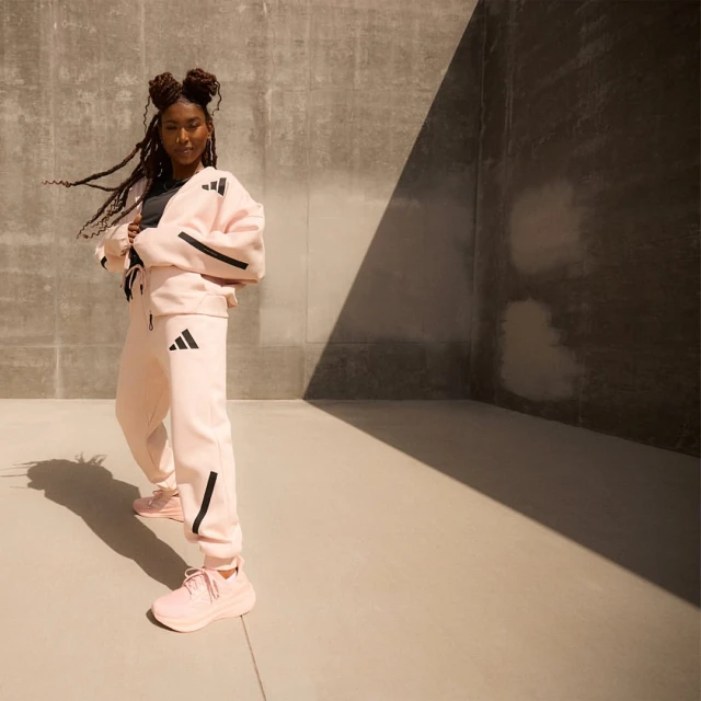 Woman in pink sweatsuit posing in a concrete corner with shadow, showcasing the Adidas logo on the jacket, pants, and running shoes.