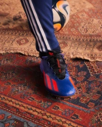 Close-up shot of a person wearing blue football boots with orange stripes and matching blue track pants standing on an ornate oriental rug.