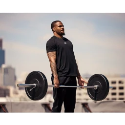 Black man holding a heavy barbell outdoors against a city skyline, wearing a black t-shirt and athletic pants.
