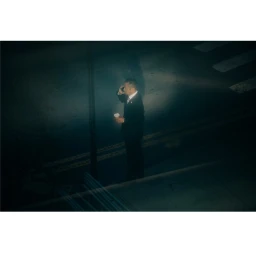 Man in a dark suit standing on a nighttime street holding a phone and a takeaway coffee cup, captured from an elevated high angle.