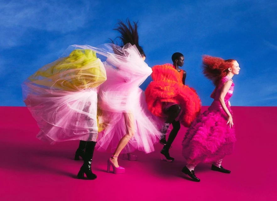 Four fashion models wearing brightly colored tulle dresses and platform shoes walk against a blue and hot pink backdrop in a studio shot.