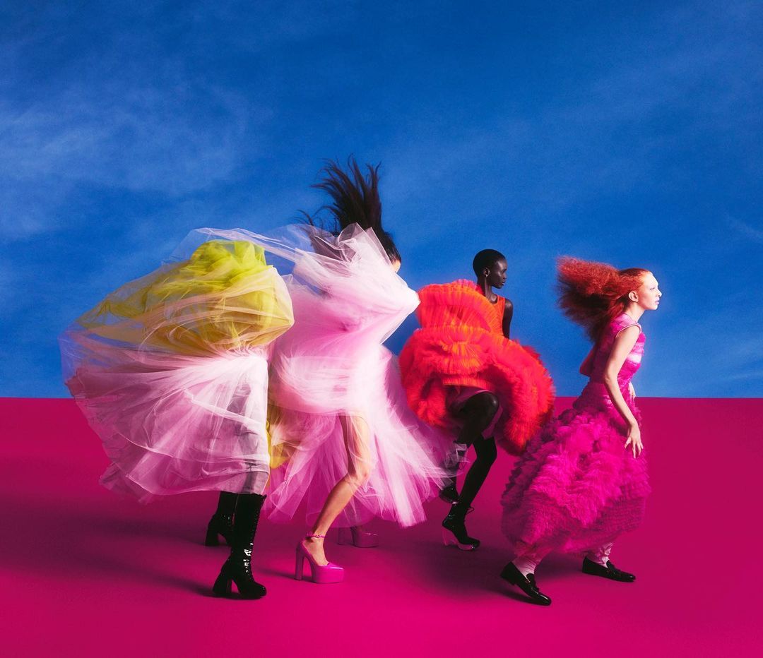 Four women in vibrant, layered tulle gowns stride across a magenta surface against a blue sky. Fashion editorial photography.