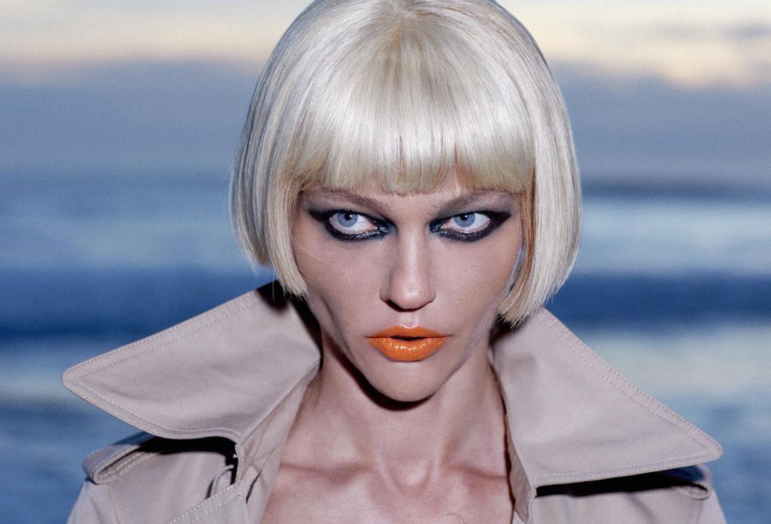 Fashion portrait of an adult female with a blonde bob haircut and heavy makeup, wearing a trench coat, posing outdoors against a blurred background.