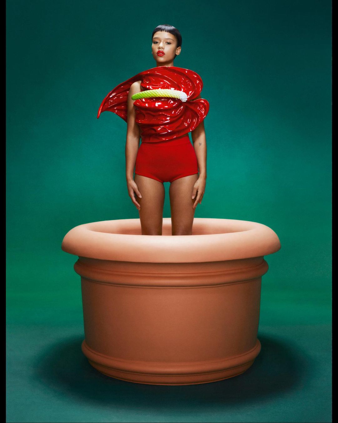 Woman in a red outfit stands inside a large terracotta flower pot against a green background in a fashion photography studio shot.