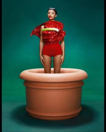 Woman in a red outfit stands inside a large terracotta flower pot against a green background in a fashion photography studio shot.