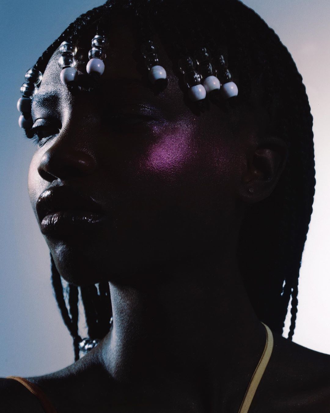 Portrait of a Black woman with braided hair and white beads, illuminated by dramatic side lighting against a gradient background.