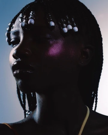 Portrait of a Black woman with braided hair and white beads, illuminated by dramatic side lighting against a gradient background.