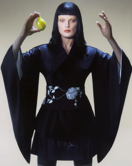 Elegant woman in a black kimono holds a yellow pear against a light beige background in a studio fashion portrait.