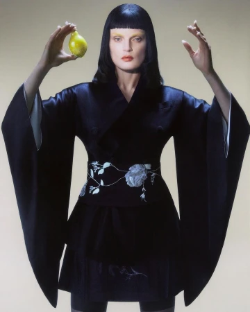 Elegant woman in a black kimono holds a yellow pear against a light beige background in a studio fashion portrait.